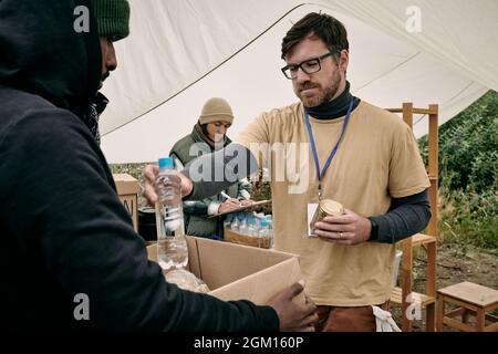 Giovane volontario bearded in vetri che tengono lattina può e che dà la bottiglia di acqua all'uomo senza tetto con scatola Foto Stock