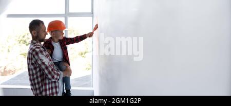 Un uomo costruttore con suo figlio in braccia sullo sfondo di una finestra. Bambino in un casco dipinge il muro bianco mentre fa le riparazioni a casa. Foto Stock