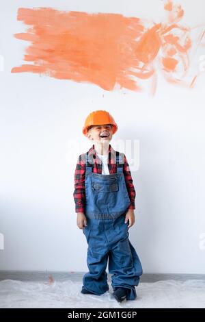 Un bambino in un casco di sicurezza e tute sullo sfondo di una parete bianca in cemento con un punto arancione per lo spazio per il testo. Foto Stock