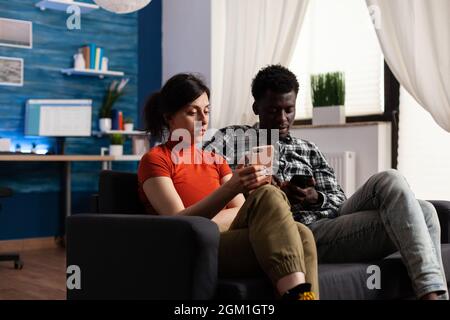 Coppia interrazziale seduta sul divano rilassante con gadget. Donna caucasica e uomo afroamericano utilizzando smartphone con tecnologia. Partner misti che detengono dispositivi telefonici. Foto Stock