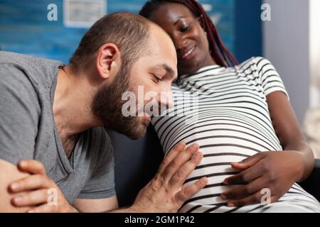 Primo piano di padre caucasico di bambino guardando il ventre e tenendo la mano mentre incinta madre nera sorridendo. Coppia interrazziale con gravidanza in attesa del bambino e rilassante a casa Foto Stock