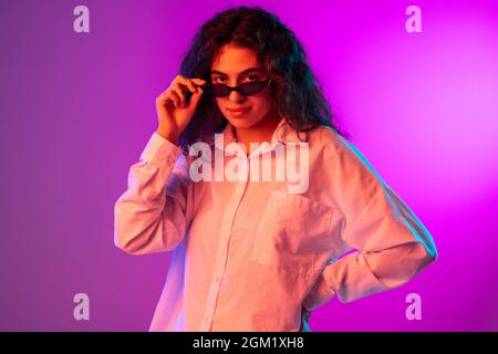 Ritratto di bella ragazza mettere occhiali da sole e guardare la fotocamera isolato su viola, lilla sfondo studio colore sfondo in luce al neon. Foto Stock