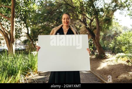 Allegra donna elegante sorridente alla macchina fotografica mentre tiene in mano una targhetta bianca. Felice donna di mezza età che mostra un banner vuoto. Standin attivista femminile Foto Stock