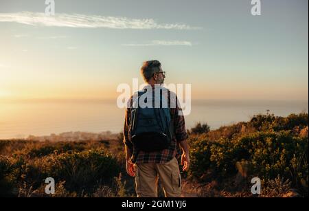 Zaino in spalla maturo che guarda la vista panoramica su una collina. Vista posteriore di un escursionista maschile in piedi da solo su una collina costiera. Avventuroso uomo maturo godei Foto Stock