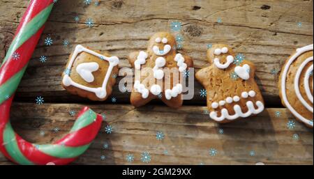 Fiocchi di neve che cadono contro i biscotti dell'uomo del pan di zenzero e la canna di caramella sulla superficie di legno Foto Stock
