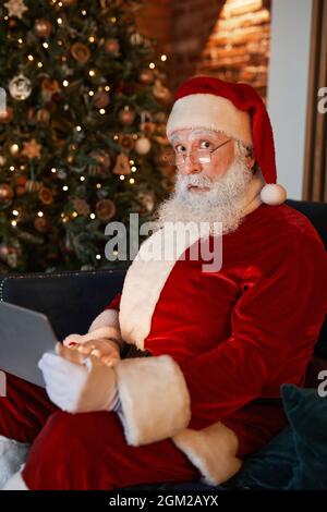 Ritratto di Babbo Natale scioccato in occhiali seduti sul divano in soggiorno con albero di Natale e utilizzando un computer portatile Foto Stock