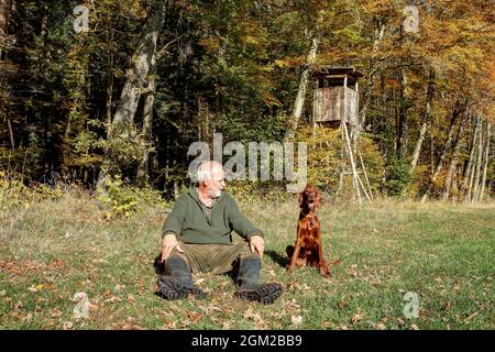 Nel pomeriggio di sole in autunno un cacciatore siede con il suo cane da caccia sul bordo colorato della foresta al sole di fronte al pulpito di caccia. Foto Stock