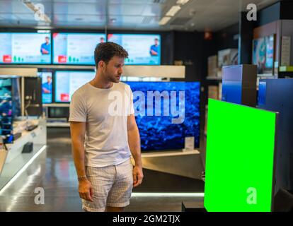 Giovane cliente maschio bearded in piedi di fronte a grande TV a schermo piatto con monitor verde chromakey mentre si visita il negozio di elettronica moderna Foto Stock