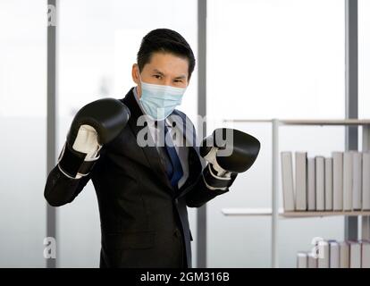 Uomo d'affari asiatico vestito di vestito e cravatta, indossando una maschera per il viso e un guanto da boxe. Postura in piedi pronta per la boxe. Il concetto di non lasciarti Foto Stock