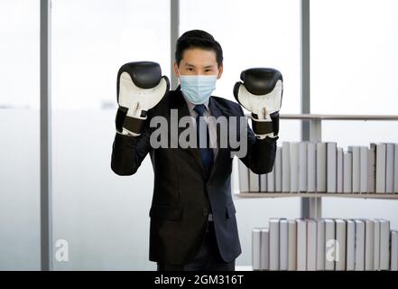 Uomo d'affari asiatico vestito di vestito e cravatta, indossando una maschera per il viso e un guanto da boxe. Postura in piedi pronta per la boxe. Il concetto di non lasciarti Foto Stock