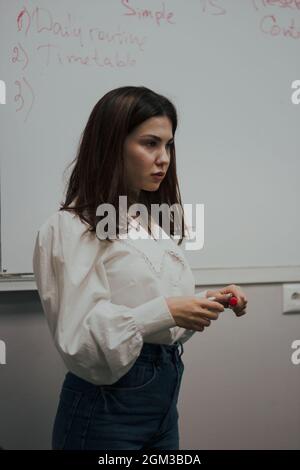 Giovane insegnante di lingua inglese femminile con un pennarello rosso in una blusa bianca in piedi di fronte a una lavagna bianca. Foto Stock