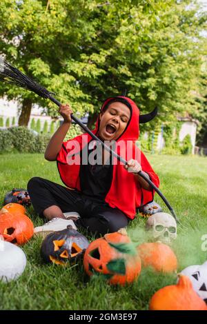 ragazzo afroamericano arrabbiato in costume diavolo di halloween urlando mentre tiene la scopa vicino alle zucche e seduto sul prato Foto Stock