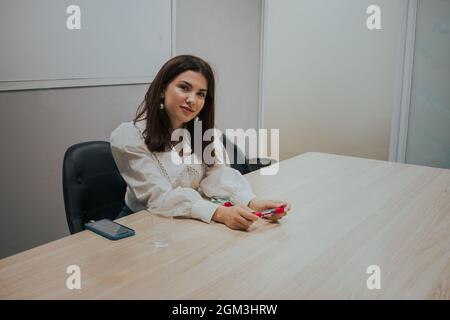 Giovane donna bruna in una blusa bianca seduta al tavolo in ufficio e tenendo un marcatore nelle sue mani. Concetto di formazione aziendale. Foto Stock