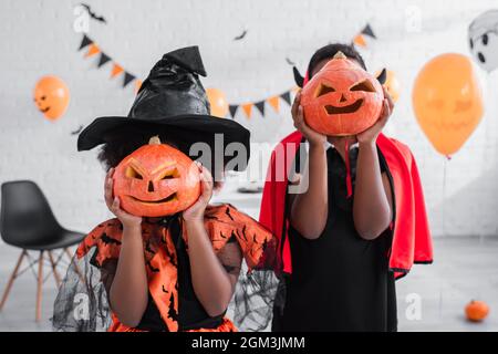 bambini afroamericani in costumi di halloween che coprono i volti con zucche scolpite a casa Foto Stock