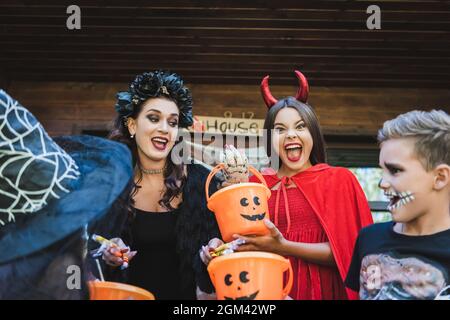 donna con caramelle vicino i capretti eccitati che tengono i secchi e grida nei costumi di halloween creepy Foto Stock