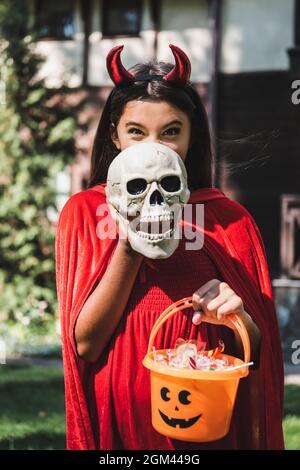 ragazzo allegro in costume diavolo di halloween faccia oscura con il cranio  spoky mentre tiene secchio di dolci Foto stock - Alamy