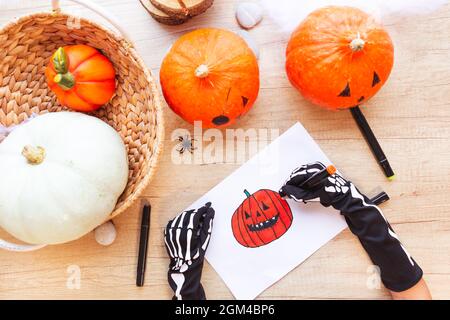 Mani in guanti neri con ossa stanno disegnando uno schizzo con marcatori una zucca ridente, su una scrivania di legno circondata da zucche e ragni, vista dall'alto Foto Stock