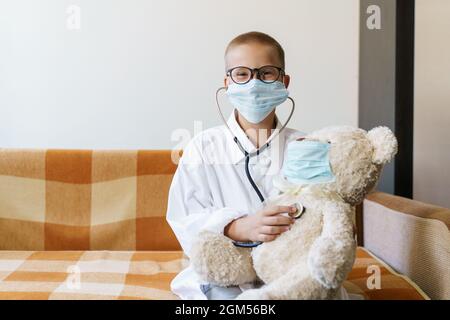 Vista del bambino che gioca medico o infermiere con orsacchiotto alla luce del sole a casa. Happy boy ascolta uno stetoscopio giocattolo. Gioco di ruolo ragazzo giocoso. In occhiali e un accappatoio con maschera protettiva Foto Stock