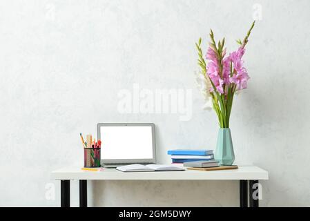 Vaso con bei fiori gladioli, moderno computer portatile, libri e cancelleria sul tavolo contro la parete di luce Foto Stock