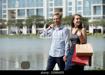 Felice giovane coppia a piedi intorno laghetto città con borse shopping dopo aver fatto acquisti in vendita Foto Stock