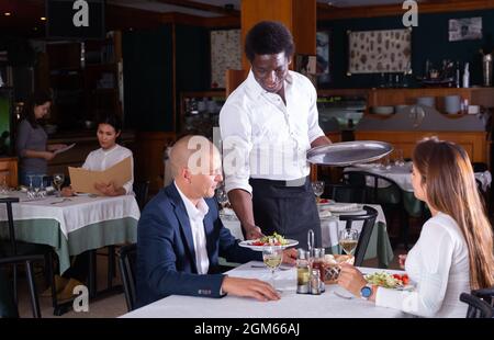 sorridente cameriere maschile portare ordine a uomo adulto e donna in restaurante Foto Stock