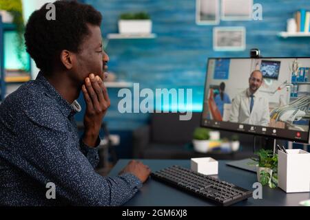 Paziente adolescente con mal di denti discutere di trattamento medico con il medico durante la videoconferenza on-line riunione. Uomo che soffre di malattie dolorose. Videoconferenza telefonica sul computer Foto Stock