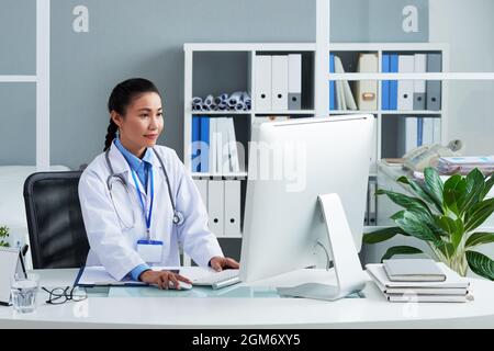 Sorridente medico femminile in labcoat lavoro sul computer nel suo ufficio e anayzing risultati di test medici Foto Stock