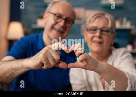 Coppia anziana che crea figura a forma di cuore con le mani a casa. Sposato persone anziane in amore facendo segno romantico mentre si guarda la macchina fotografica in soggiorno. Marito e moglie mostrano gesto di affetto Foto Stock