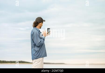 L'uomo caucasico con sta tenendo uno smartphone in mano e prendendo un selfie su una spiaggia. Foto Stock