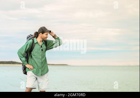 Il viaggiatore con il suo zaino turistico è in piedi su uno sfondo marino. L'uomo sta togliendo il suo cappellino da baseball e guardando a distanza. Foto Stock