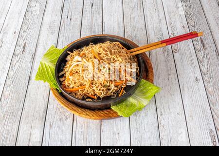 Spaghetti cinesi ricci fritti con verdure fritte e recipiente in metallo con buco di pollo e bacchette sul tavolo bianco Foto Stock