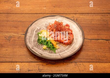 Tartare di tonno rosso e cocomero con semi di sesamo e papavero. Carota grattugiata, insalata di wakame, mango tritato e filamenti di zafferano su piatto rotondo e legno Foto Stock