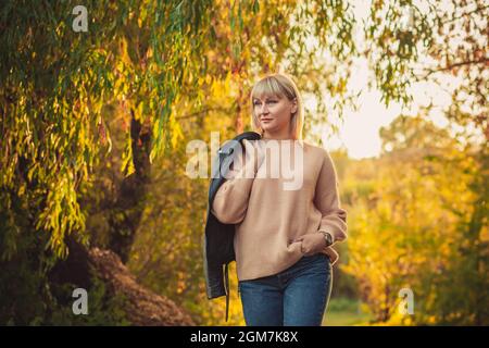 Una donna bionda con un taglio corto cammina attraverso la foresta in un maglione a maglia e una giacca in pelle sulla spalla. Escursioni all'aperto in autunno fores Foto Stock