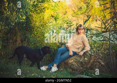 Una donna bionda in un maglione lavorato a maglia con un taglio corto è seduta su un albero caduto e un cane nero rottweiler corre intorno nella foresta. Passeggiata nella natura Foto Stock