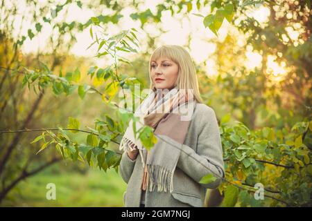 Una donna bionda con un taglio corto cammina attraverso la foresta in un cappotto di lana grigio e una sciarpa di plaid. Passeggiate nella natura in autunno foresta Foto Stock