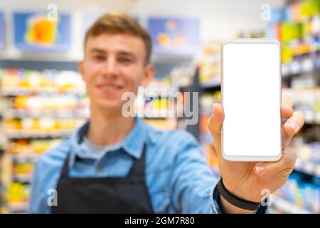 Foto sfocata di un uomo sorridente negozio che indossa grembiule nero e orologio digitale in mano che mostra lo smartphone bianco schermo di colore bianco Foto Stock