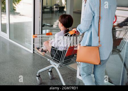 Back view photo di una ragazza di età scolastica seduta nel carrello della drogheria e sua madre spinge il carrello per entrare nel negozio. Foto Stock