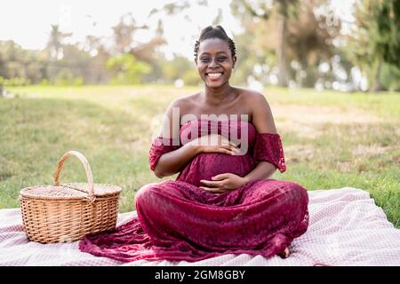 Donna africana carezzando il suo ventre incinta mentre fa un picnic nel parco - concetto di stile di vita di maternità Foto Stock