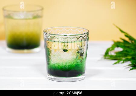 Chlorophyll verde o Chlorella bere in vetro con acqua su tavola bianca, sfondo arancione. Gocce verdi in acqua limpida, cibo antiossidante e bevande. Foto Stock