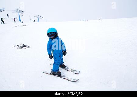 il bambino in casco, occhiali, sci e tute blu si alza sul fianco nevoso della montagna. Lezione di sci per bambini presso la scuola di sci. Animazione invernale attiva f Foto Stock