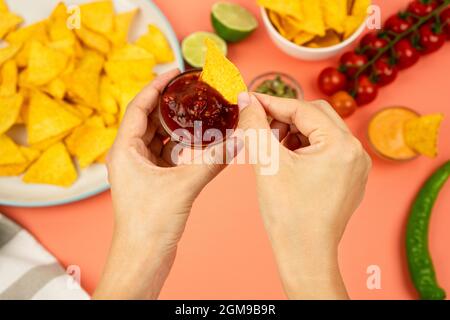 Mani che immergere nachos in salsa di pomodoro sullo sfondo di cibo e verdure messicane. Foto Stock