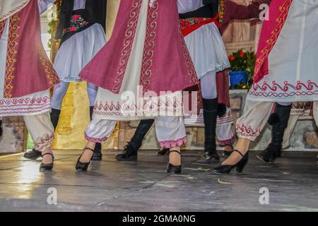 Vista ritagliata dei ballerini greci sul palco in bellissimo costume ricamato con le donne di fronte agli uomini - gambe e piedi - movimento sfocato Foto Stock