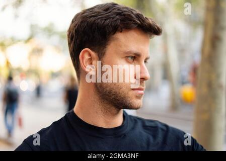 Serio e pensivo giovane uomo con pettinature eleganti vestito in abito casual in piedi su sfondo urbano sfocato e guardando via Foto Stock