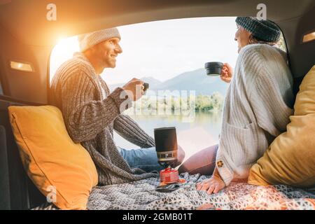 Vista bagagliaio auto di coppia chatting vestiti caldi a maglia godendo di gas stufa preparato caffè e vista lago di montagna. Accogliente coppia di inizio autunno aut Foto Stock