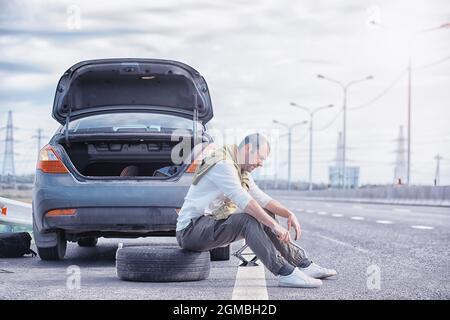 Sostituzione della ruota di una vettura su strada. Un uomo che fa il lavoro di pneumatico ai margini. Foto Stock