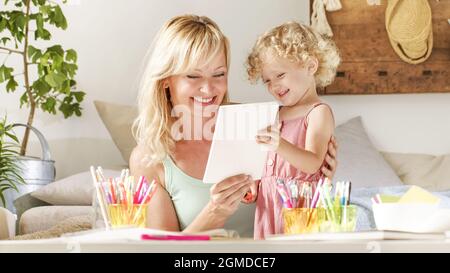 sorridendo la madre sorride a bambino biondo ricci capelli, attività di apprendimento prescolare a casa, concetto di crescita sana, disegna e scrive cheerfu Foto Stock