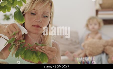 La donna bionda si prende cura delle piante nel soggiorno, spruzza sulle foglie verdi mentre la sua figlia bambino guarda la sua curiosamente, casa hobby conce Foto Stock