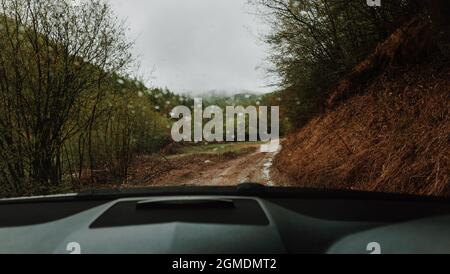 Punto di vista interno del parabrezza dell'auto POV. Guida di un veicolo fuoristrada moderno con guida a destra sulla strada di campagna sporca della foresta di montagna. Riflessione della faccia Foto Stock