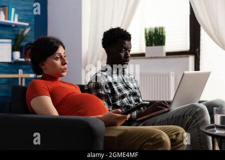 Coppia interrazziale in attesa di bambino e utilizzando la tecnologia a casa. Uomo afroamericano che lavora su un moderno computer portatile mentre incinta donna caucasica che cambia canale con telecomando TV. Foto Stock