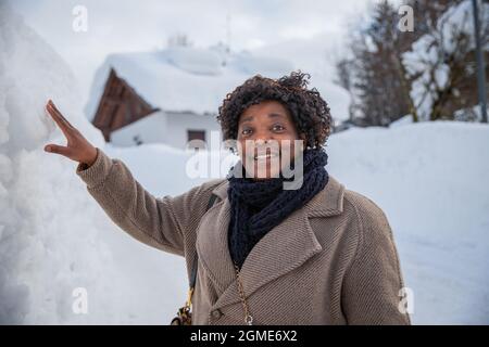 Ritratto di una donna africana sorridente di mezza età in montagna in inverno con un sacco di neve, indossa un cappotto e sciarpa Foto Stock